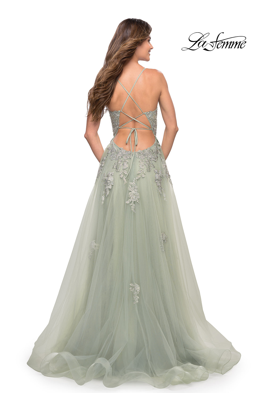 A woman wears a light green La Femme Open-Back Long Ball Gown with lace, featuring floral embroidery, a crisscross open back, and a full tulle skirt. The La Femme brand appears in the top right corner.